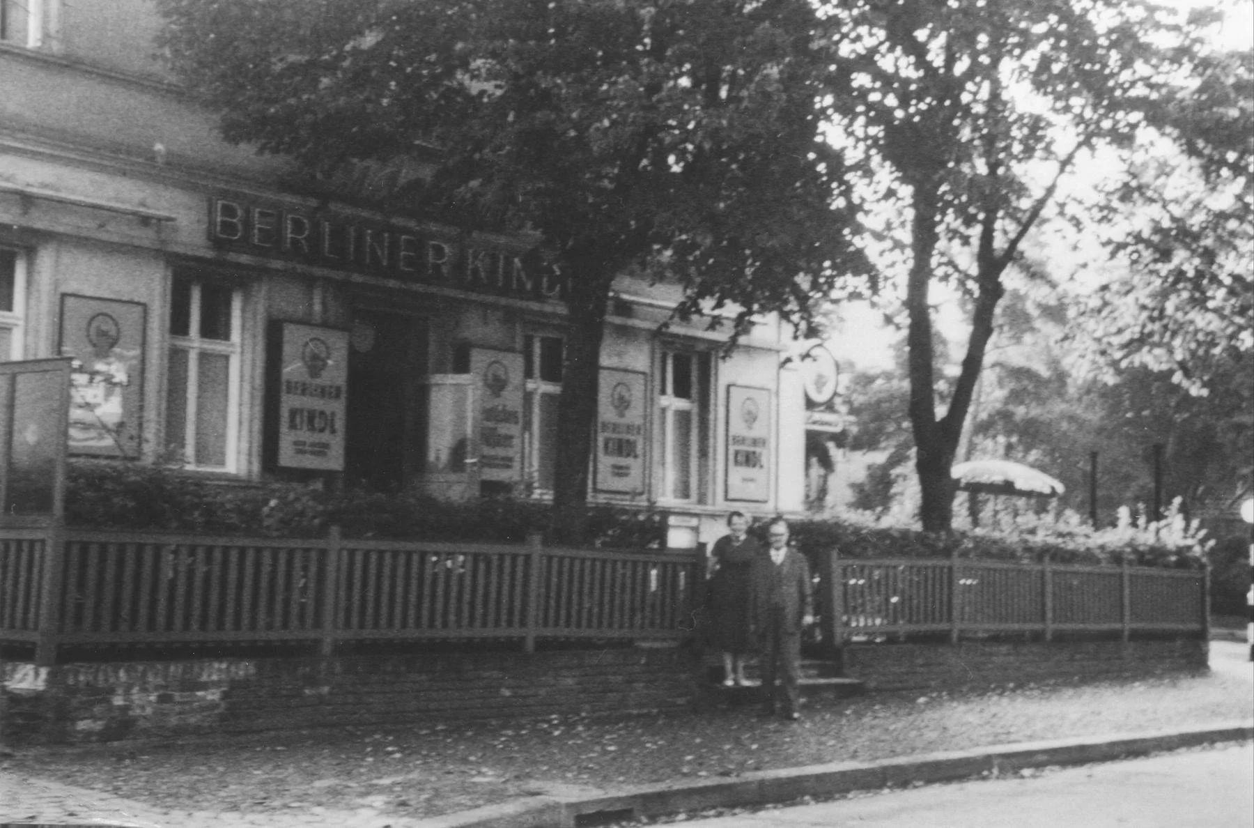 Historisches Foto aus den 1920er-Jahren: Eckhaus mit Schriftzug Berliner Kindl, Holzzaun und zwei Personen vor dem Eingang, schwarzweiß